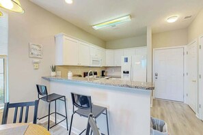 A modern kitchen with white cabinets, a granite countertop, and a central island with bar stools. - Shoreline Serenity at River Oaks (Myrtle Beach)