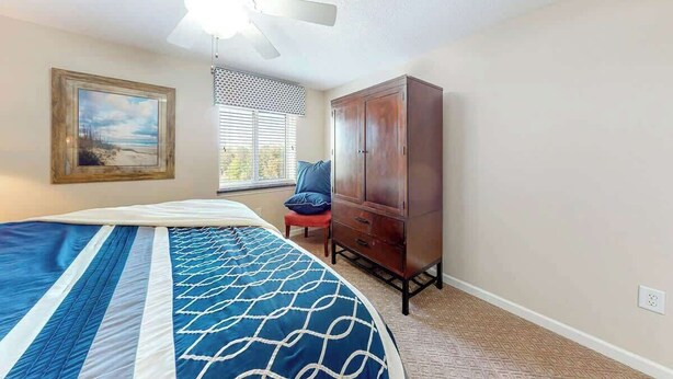 A bedroom with a bed, a wardrobe, a chair, a window with blinds, and a ceiling fan. - Yacht Club Villas 3-505 (North Myrtle Beach)
