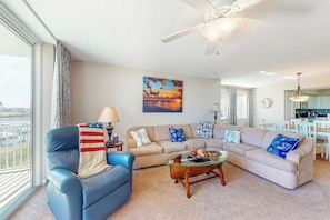 A living room with a blue armchair, a beige sofa, a coffee table, and a painting of a sunset. - Yacht Club Villas 3-505 (North Myrtle Beach)