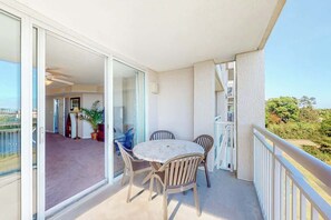 An open balcony with a table and chairs, a sliding glass door leading to an interior space, and a view of greenery.