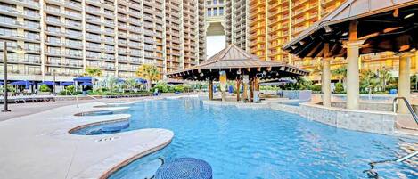 A hotel pool area with multiple swimming pools, a gazebo, and high-rise buildings in the background.
