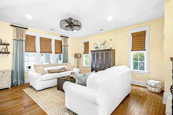 A living room with a white sofa, a coffee table, and a cabinet. There are windows with blinds and a ceiling fan. - Barefoot Livin' Retreat (North Myrtle Beach)