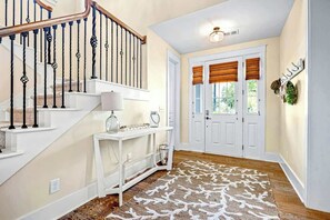 A staircase with a wooden handrail and black balusters, a white console table with a lamp, a patterned rug, and a double door with a window. - Barefoot Livin' Retreat (North Myrtle Beach)