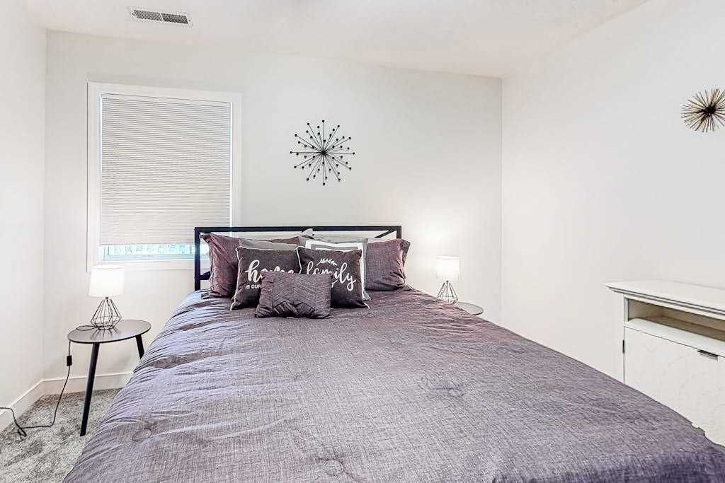 A bedroom with a bed, a bedside table, a lamp, a wall clock, and a window with blinds.