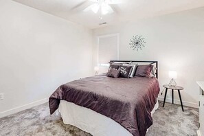 A bedroom with a bed, a bedside table, a wall clock, and a window with blinds.
