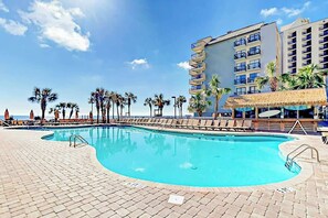 A swimming pool with lounge chairs and umbrellas, surrounded by palm trees and a hotel building. - Cast a Wave, Unit 352 (Myrtle Beach)