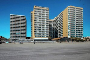 Ein modernes Hochhaus mit mehreren Balkonen, einer flachen Dachterrasse und blauem Himmel.