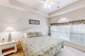 Ein Schlafzimmer mit einem Bett, einem Nachttisch, einer Lampe, einem Deckenventilator, einem Fenster mit Jalousien und einem Wandbild.