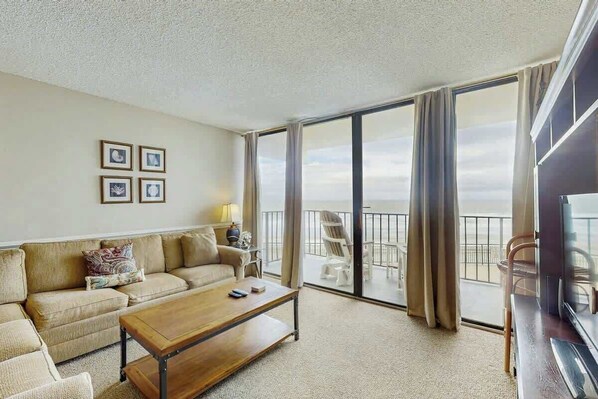 Habitación de hotel con sofá, una mesa de centro, televisor y balcón con vistas.