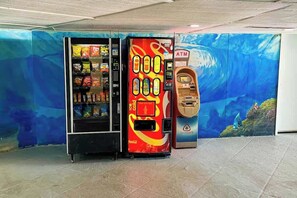 Ein Automat mit tropischem Wandbild im Hintergrund, ein Geldautomat und ein Verkaufsautomat für Snacks.