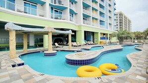 Zona de piscina de hotel con varias piscinas, sillas de descanso y un edificio con balcones.