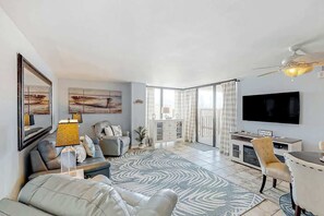 A modern living room with a sofa, a coffee table, a TV, and a large window. - Sea Castle Unit A-3 Condo (North Myrtle Beach)