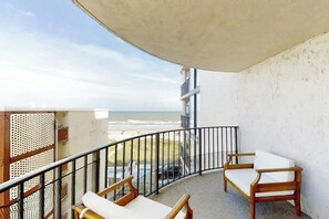 Een balkon met uitzicht op het strand, een houten stoel, een klein tafeltje en een witte muur.