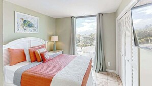 Ein Schlafzimmer mit Bett, Nachttisch, Lampe, einem Fenster mit Vorhängen und Blick ins Freie.