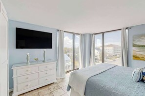 Een slaapkamer met een bed, een dressoir, een flatscreen televisie en grote ramen met uitzicht op gebouwen en een landschap.