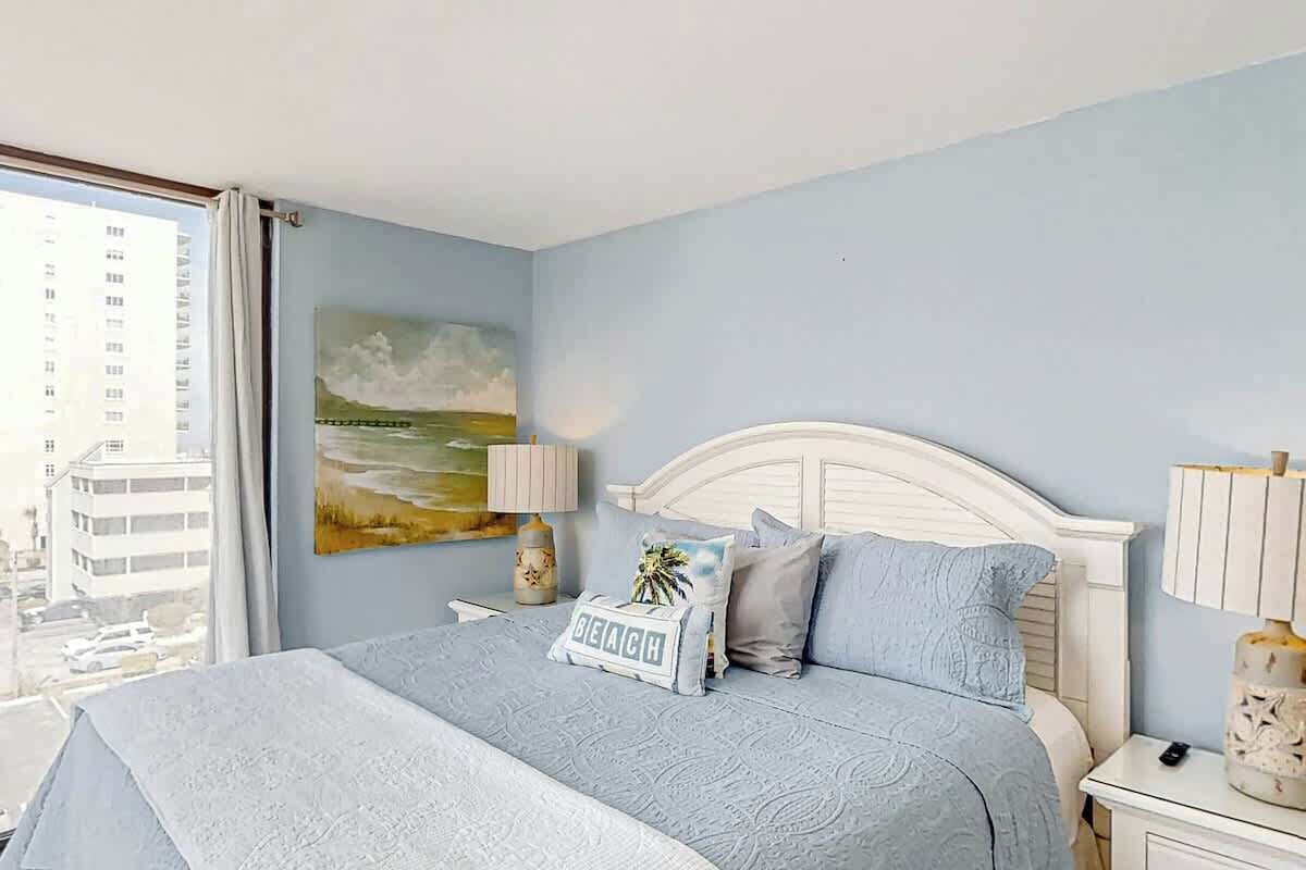 A hotel room with a bed, a nightstand, a lamp, a painting, and a view of buildings outside.