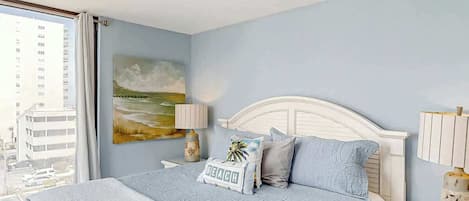 A hotel room with a bed, a nightstand, a lamp, a painting, and a view of buildings outside.
