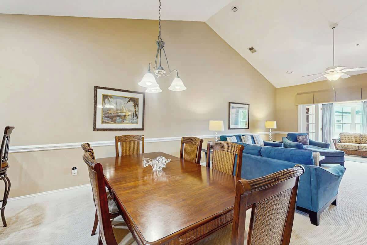 A dining area with a wooden table and chairs, a chandelier, and a living room with sofas and a ceiling fan.