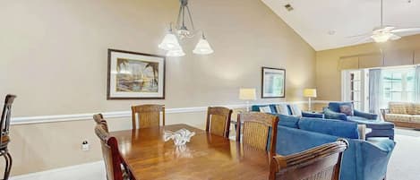 A dining area with a wooden table and chairs, a chandelier, and a living room with sofas and a ceiling fan.