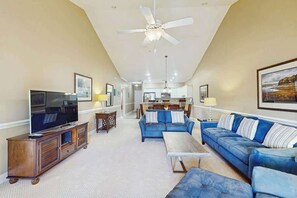 Ein geräumiges Wohnzimmer mit einem Flachbildfernseher, blauen Sofas, einem Couchtisch und einem Deckenventilator.