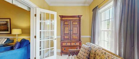 A room with a blue sofa, a patterned sofa, a wooden cabinet, and a large window with curtains.