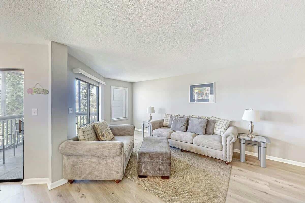 A living room with a grey sofa set, a coffee table, and a rug. There are two lamps on side tables, and a picture frame on the wall.