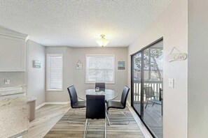 A dining area with a glass table and chairs, a kitchen with white cabinets, and a view through a glass door to an outdoor deck. - Ocean Greens Unit D-402 (North Myrtle Beach)