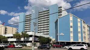 A hotel with multiple floors and balconies, a parking lot with several cars, and a clear sky with clouds. - Water's Edge Resort 1002 (Murrells Inlet)
