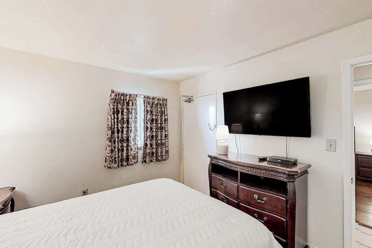 Un dormitorio con cama, televisor de pantalla plana en la pared, cómoda y ventana con cortinas estampadas.