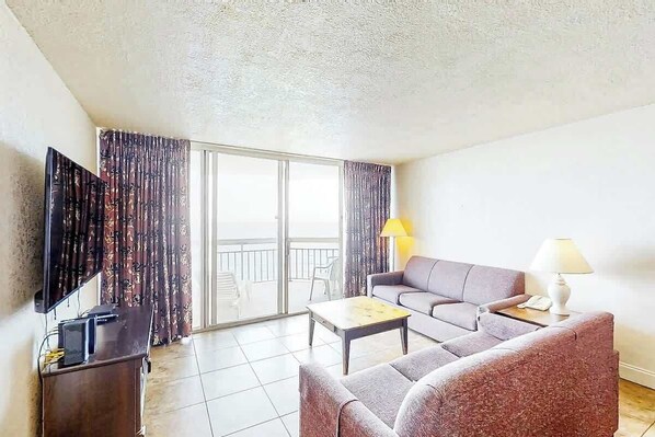 Un salon avec un canapé, une table basse, un téléviseur et un balcon agrémenté de rideaux.