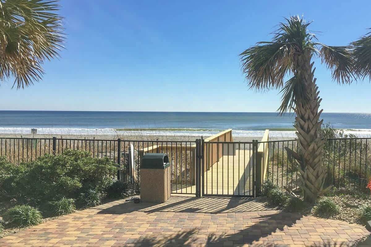 Un chemin pavé mène à une plage, avec une barrière en métal, des palmiers et une vue dégagée sur l’océan.