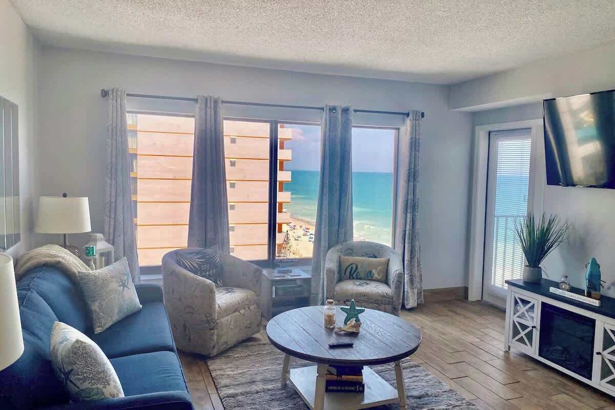 A living room with a sofa, armchairs, a coffee table, and a TV. There are large windows with a view of the beach.