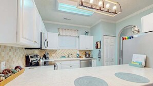 A modern kitchen with white cabinets, a central island with a built-in induction cooktop, and a refrigerator with a colorful magnet strip. - Seas the Day at Magnolia Place (Myrtle Beach)