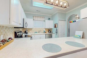 A modern kitchen with white cabinets, a central island with a built-in induction cooktop, and a refrigerator with a colorful magnet strip. - Seas the Day at Magnolia Place (Myrtle Beach)