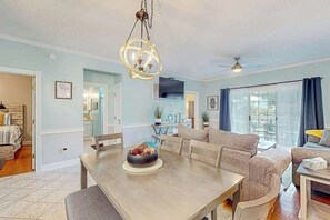 A spacious living room with a dining area, a ceiling fan, and a large window with curtains. - Seas the Day at Magnolia Place (Myrtle Beach)