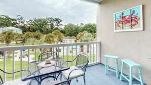 A balcony with a table and chairs, a painting on the wall, and a view of a garden and houses. - Seas the Day at Magnolia Place (Myrtle Beach)
