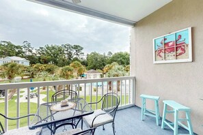 A balcony with a table and chairs, a painting on the wall, and a view of a garden and houses. - Seas the Day at Magnolia Place (Myrtle Beach)