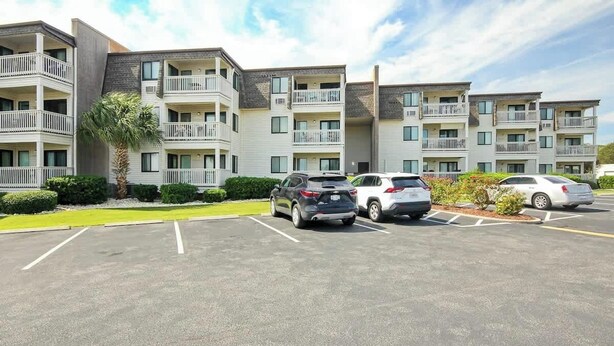 A multi-story parking structure with several cars parked, surrounded by a landscaped area with a palm tree. - Ocean Forest Villas E-308 (Myrtle Beach)