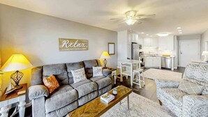 A living room with a grey sofa, a wooden coffee table, and a kitchen in the background. - Ocean Forest Villas E-308 (Myrtle Beach)