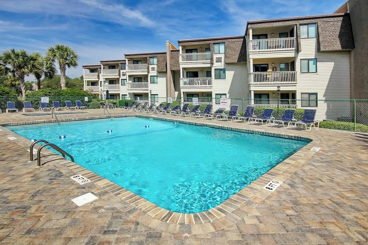 Una piscina rectangular con solárium empedrado, rodeada de sillones de descanso y un edificio de varios pisos con balcones.