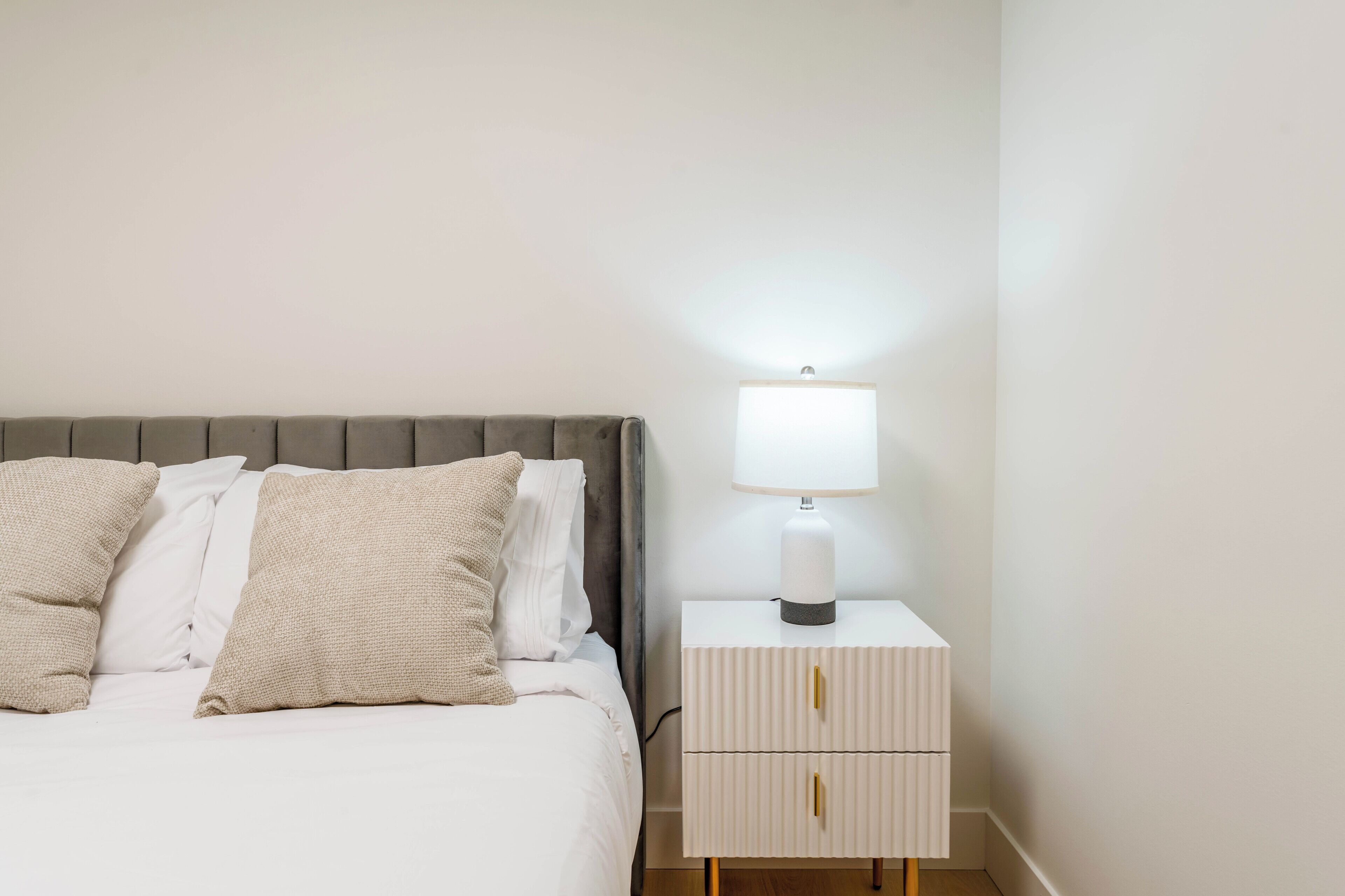 En pænt redt seng med gråt sengegavl og hvide sengetæppe, et natbord med en lampe og en lille skuffeunit.