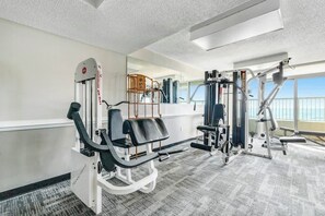 A gym with various exercise equipment, a mirror, and a view of the outside through large windows. - Camelot by the Sea 0303 (Myrtle Beach)
