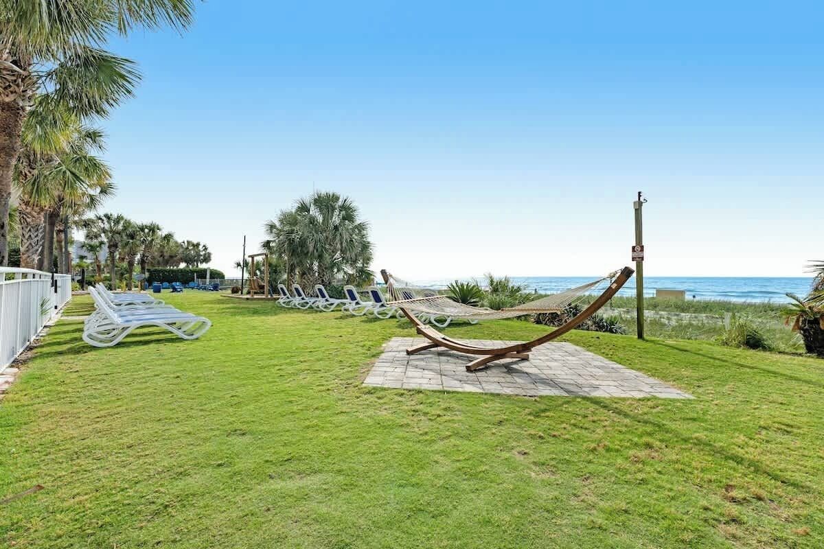 Eine Rasenfläche mit einer Hängematte und Liegestühlen, einem gepflasterten Weg und Blick auf das Meer.