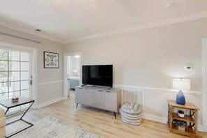 A living room with a flat-screen TV on a grey cabinet, a small round ottoman, a white rug, and a side table with a lamp and vase. - Magnolia Escape Retreat (Myrtle Beach)