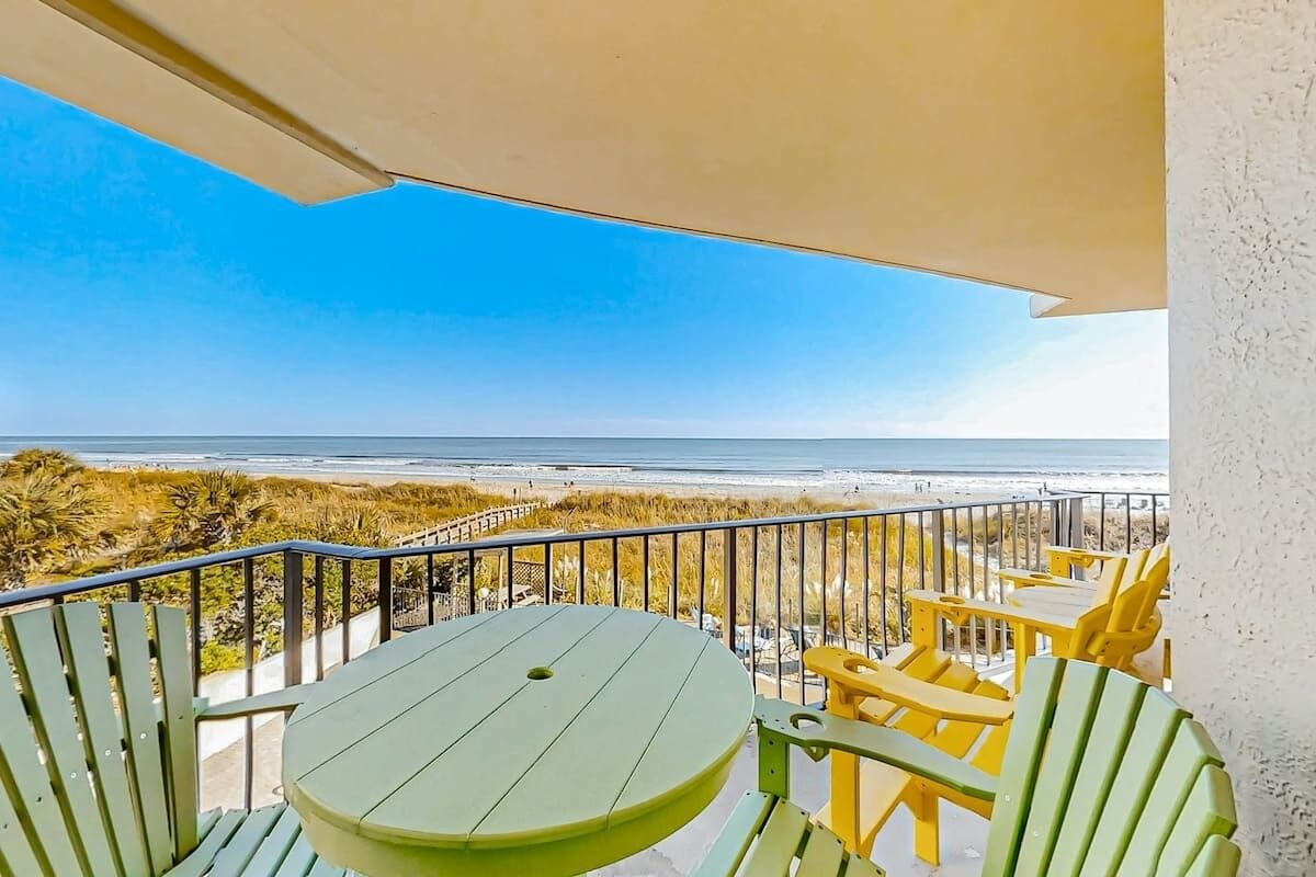 A balcony with a table and chairs, overlooking a beach and ocean.