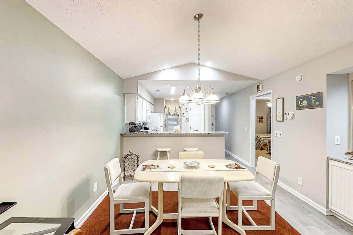 A dining area with a white table and chairs, a chandelier, and a kitchen in the background.