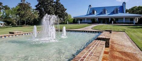A clear pool with a fountain, surrounded by a brick deck, and a large blue building in the background.