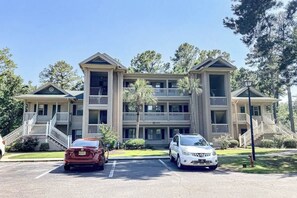 A multi-story building with a central entrance, balconies, and a parking area with two cars. - Pinehurst Condos Unit 6J (Pawleys Island)