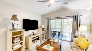 Una sala de estar con televisor de pantalla plana, estanterías de madera, una mesa de centro con adornos, un sofá con cojines amarillos y un ventilador de techo.