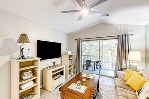 Ein Wohnzimmer mit Flachbildfernseher, Holzregalen, einem Couchtisch mit Dekoration, einer gelben Kissen ausgelegten Couch und einem Deckenventilator.
